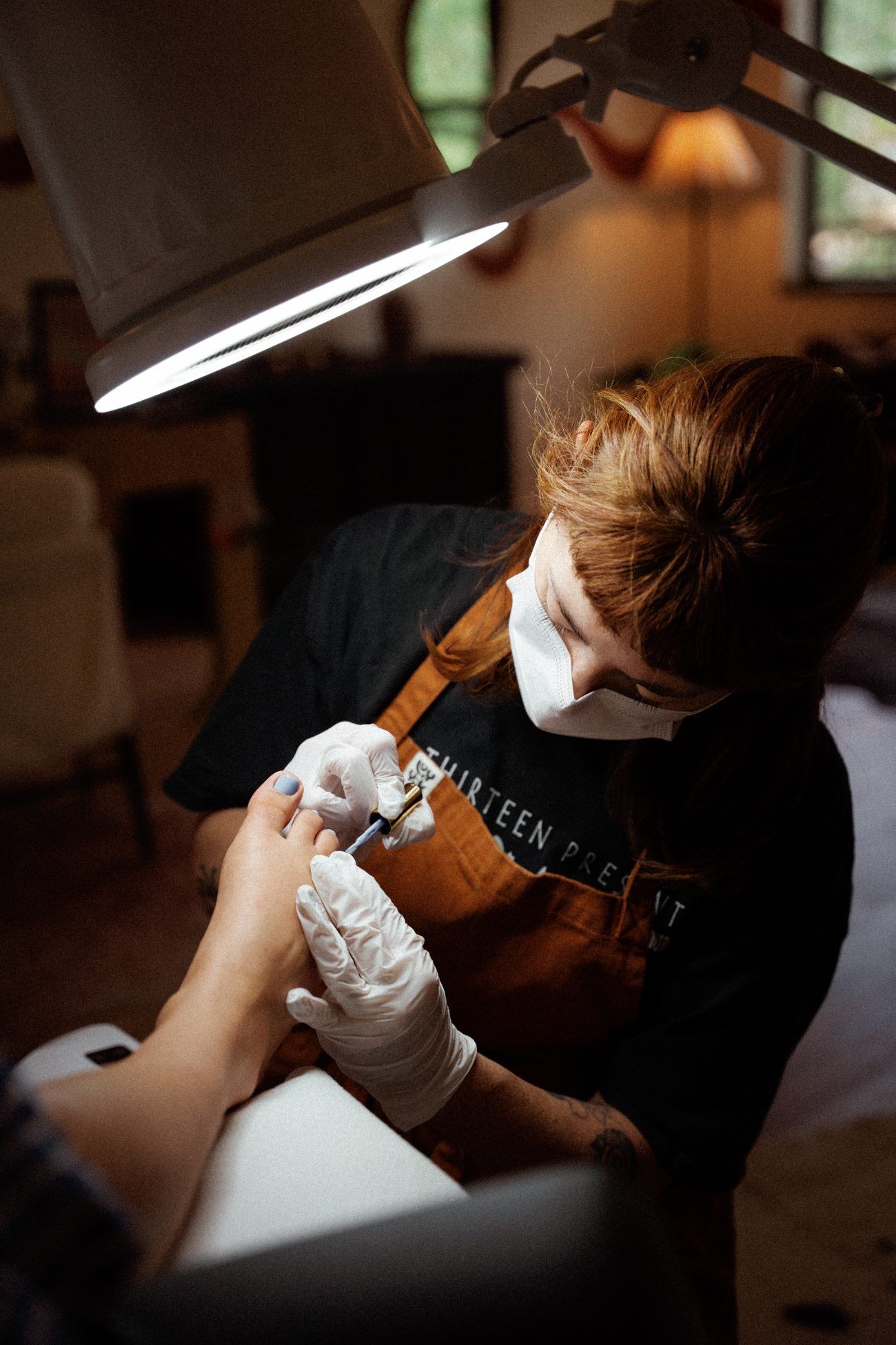 Kira in mask and gloves painting a client's toenails periwinkle under the magnifying studio lamp